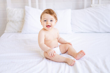 a joyful baby girl of six months on a bed in diapers smiles, a small child on a cotton bed at home, the concept of care and hygiene