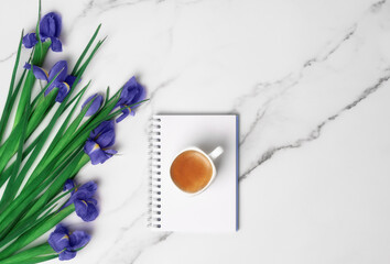 Cup of hot morning coffee on notepad and violet irises on marble background. Flat lay with copy space for text. Greeting card for Women's Day and March 8th.