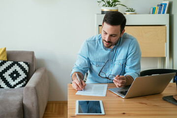 Working at Home Office: Serious  Business Man Sitting at Table with Laptop Computer, Digital Tablet and Signing a Contract or Doing Paperwork in the Living Room