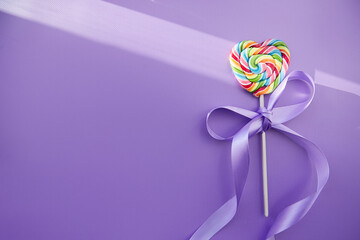 Rainbow heart shaped lollipop sweet on purple background with festive ribbon and shadows. Copy space. Minimalist concept