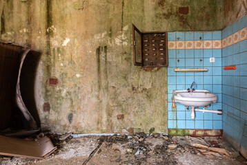 Old abandoned and run-down room in a hotel with sink against blue tiles and a destroyed closet among clutter