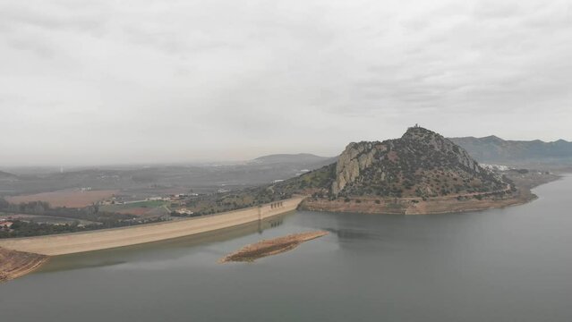 Aerial images showing a huge reservoir from 120 meters high.  In the background a large mountain with a medieval castle on it.  Camera is approaching to the dam. A little island in the foreground