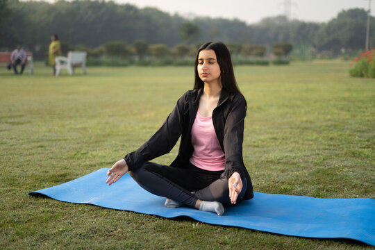 Young indian girl doing yoga on a yoga mat in a green park, early in the morning. Girl wearing black yoga pants. Healthy lifestyle and yoga concept.