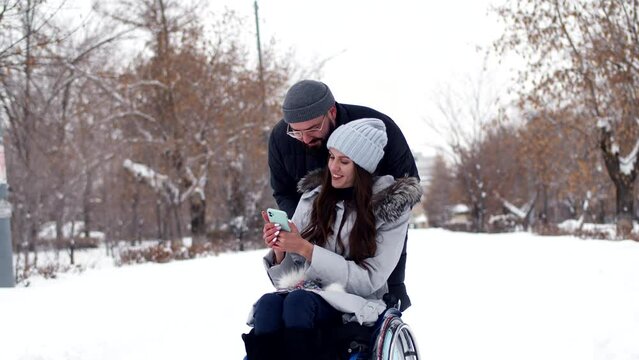Man is pushing wheelchair through winter park in which woman is disabled, they are talking looking at smartphone, front view. Disabled woman spends time with man in winter park