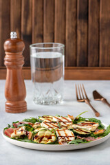 Light salad of young zucchini, bacon, halloumi cheese with arugula. A glass of water is on the table.