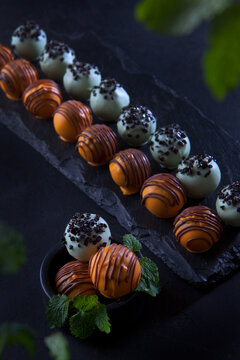 Handmade round shaped candies. A black plate made of stone. Belgian chocolate of the highest quality. Mint leaves are out of focus. Dessert for the holiday. Black background.Vertical photo.