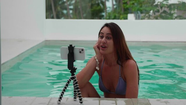 Smiling young woman recording video blog podcast on smartphone on a tripod in pool. Female blogger shooting mobile vlog, live stream, calling online concept