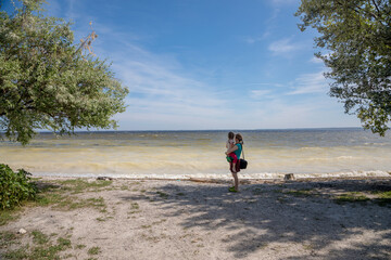 A mother with a child in her arms looks at the water from a small bay on the shore, overgrown with trees and bushes.