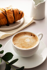 Coffee cup with fluffy milk foam and croissantson bright background. Breakfast time