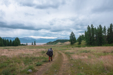 Fototapeta na wymiar Tourist walks through hills and forest towards bad weather. Hiker on way to large snow mountain range under rainy cloudy sky. Man in raincoat in mountains in overcast. Traveler goes towards adventure.