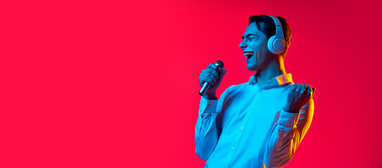 Portrait of young cheerful man in white headphones singing in microphone isolated over red studio background in blue neon