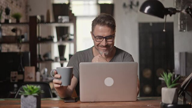 Bearded man working online with laptop computer at home sitting at desk. Home office, browsing internet. Portrait of mature age, middle age, mid adult man in 50s.