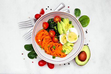 Healthy breakfast bowl with sweet potato, egg, avocado and spinach. Above view with frame of ingredients on a white marble background.