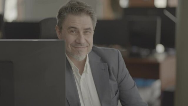 Business portrait - confident businessman sitting at desk in office. Happy mid adult man in shirt and jacket, smiling. Bearded, gray hair. Video in LOG format - S-LOG3 Cine.