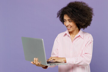 Fototapeta na wymiar Young smiling happy cheerful fun woman of African American ethnicity 20s wear pink striped shirt hold use work on laptop pc computer isolated on plain pastel light purple background studio portrait.