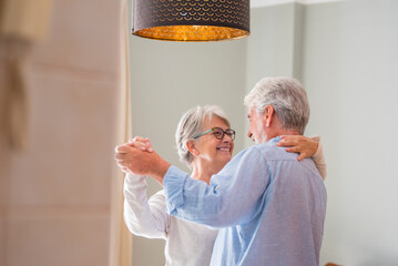 Romantic loving senior couple holding hands enjoying dancing together in the living room of house,...