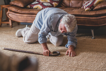 Elder senior man lying on floor after falling down with wooden walking stick beside couch on rug in living room at home. Old man suffering with pain and struggling to get up after falling down  - Powered by Adobe