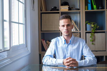 Portrait of handsome young business executive sitting on desk with hands clasped and looking at camera. Confident caucasian businessman in formal shirt in front of office storage cabinet