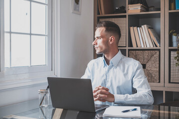 Serious young businessman sitting at office and looking out through window thinking about future career or success. Thoughtful caucasian guy sitting at desk with laptop while looking away and admiring