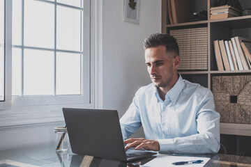 Portrait of handsome businessman typing on laptop keyboard at office. Young caucasian male executive doing online work at home. Focused man entrepreneur at work typing on keypad with document 