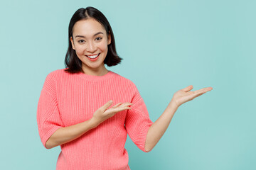 Young smiling happy woman of Asian ethnicity 20s wearing pink sweater hands arms finger aside on workspace area mock up isolated on pastel plain light blue background studio. People lifestyle concept. - obrazy, fototapety, plakaty
