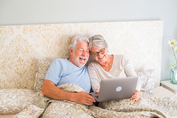 Cheerful senior couple browsing and watching social media content using laptop in bed at home. Relaxed old husband and wife spending leisure time in bed, sharing online movie in bedroom at apartment