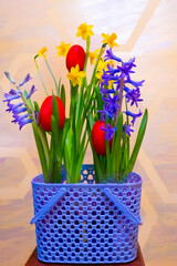 a very peri basket with spring flowers - yellow daffodils and purple hyacinths, and red painted Easter eggs against a yellow wall with hexagons.
