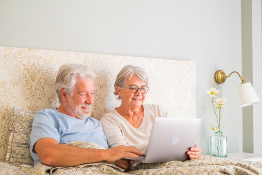 Smiling senior couple browsing and watching social media content using laptop in bed at home. Relaxed old husband and wife spending leisure time in bed, sharing online movie in bedroom at apartment