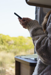 Vertical image of a woman using her smart phone inside a camper van, with the door open to enjoy a magnificent day.