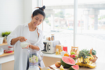 An Asian woman is making a fruit milk smoothie in her kitchen with ingredients such as watermelon,...