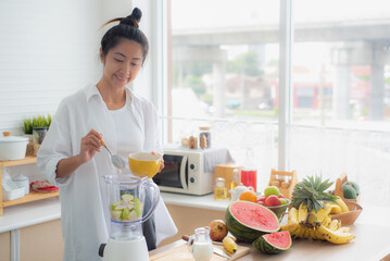 An Asian woman is making a fruit yogurt smoothie in her kitchen with ingredients such as...