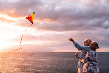 Cheerful senior couple having fun flying kite in front of beautiful seascape against cloudy sky....