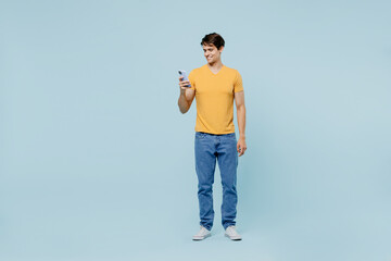 Fototapeta na wymiar Full body young man 20s wearing yellow t-shirt hold in hand use mobile cell phone browsing surfing internet isolated on plain pastel light blue background studio portrait. People lifestyle concept