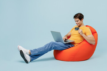 Full body young man in yellow t-shirt sit in bag chair using laptop pc computer hold credit bank...