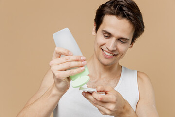 Attractive young man 20s perfect skin in undershirt hold in hand bottle tonic apply lotion on cotton pad isolated on pastel pastel beige background. Skin care healthcare cosmetic procedures concept.