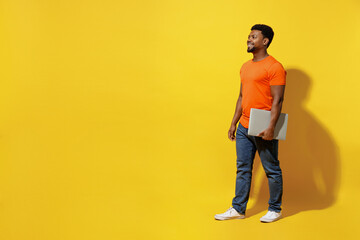 Fototapeta na wymiar Full body student fun young man of African American ethnicity 20s wear orange t-shirt hold closed laptop pc computer walk isolated on plain yellow background studio portrait. People lifestyle concept.