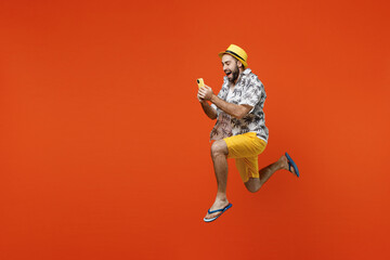 Fototapeta na wymiar Full body side view young tourist man wear beach shirt hat jump high run fast use hold mobile cell phone isolated on plain orange background studio portrait. Summer vacation sea rest sun tan concept.