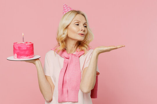 Elderly happy woman 50s wear t-shirt birthday hat hold cake show empty palm with empty place workspace area isolated on plain pastel pink background studio portrait. Celebrating party holiday concept. - Powered by Adobe