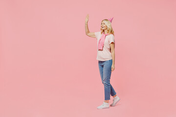 Full body elderly smiling fun cool happy woman 50s wear t-shirt birthday hat walk go waving hand say hello isolated on plain pastel pink background studio portrait. Celebrating party holiday concept.