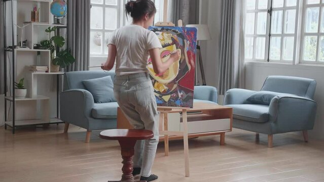 Hind View Of An Asian Artist Girl Coming To Sit On Round Wooden Chair Without Backrest And Holding Paintbrush Mixed Colour Before Painting A Girl On The Canvas
