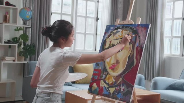 Medium Shot Side View Of An Asian Artist Girl Holding Paintbrush Mixed Colour And Painting A Girl'S Ear On The Canvas

