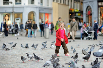 Cute little girl feeding and chasing birds on Dam Square in Amsterdam on summer day.