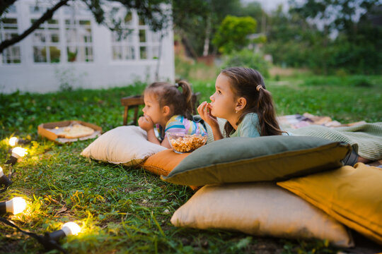 Family summer outdoor movie night. Girls lying on blanket and pillows, eating homemade popcorn and watching film on DIY screen with from projector. Summer outdoor weekend activities with kids.