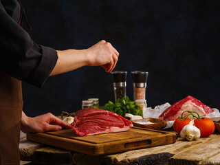 A professional chef prepares a large piece of meat - beef, pork, veal on a wooden cutting board on a dark blue background. Vegetables, spices, herbs. Hotel, restaurant, cookbook.