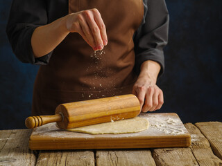 The chef rolls out floured dough on a wooden table with a rolling pin. Levitation. Dark background. Recipes for dough products - pizza, pasta, confectionery.