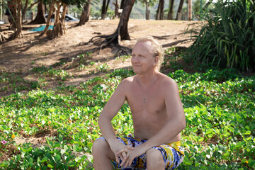 Naklejka na ściany i meble Mature blond man in shorts sits on green tropical grass on the coast, looks into the distance and smiles. Travel and tourism