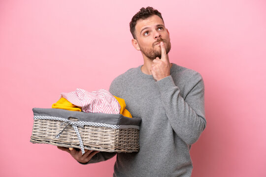 Young Brazilian man holding a clothes basket isolated on pink background having doubts while looking up