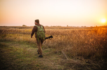 Young strong soldier with riffle and ammunition belt moving outdoors by the dirt road in sunset time