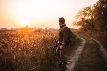 Young strong soldier with riffle and ammunition belt moving outdoors by the dirt road in sunset time