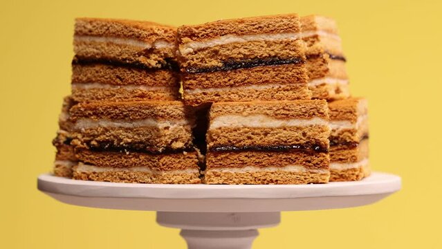 Traditional homemade Romanian Honey Layered Cake with plum jam and semolina cream also called ''Albinita'' or "Claudia". Cut portions of cake are on a cake stand, rotating against a yellow background.